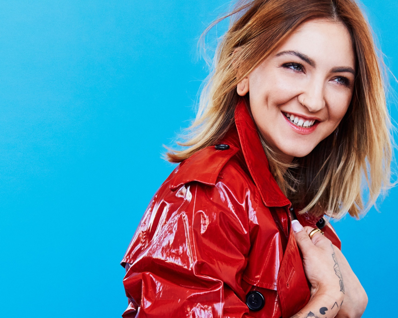 Улыбающаяся певица Джулия Майклз в красной куртке на голубом фоне