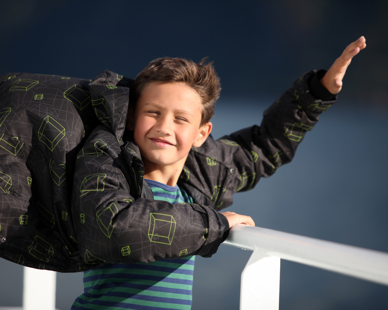 Веселый мальчик в куртке стоит на ветру в лучах солнца