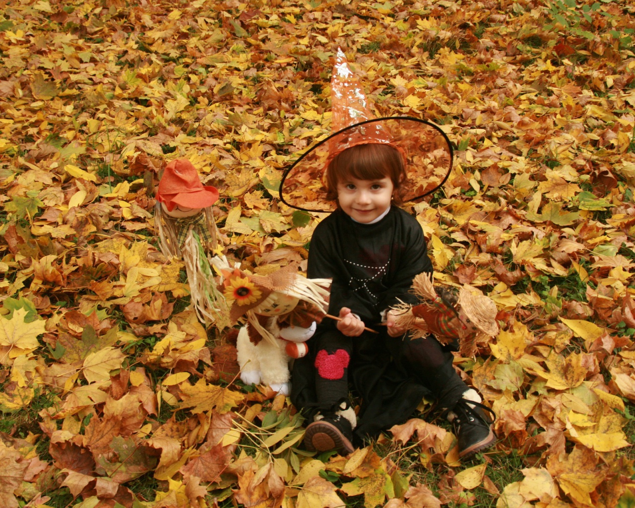 Маленькая девочка в костюме ведьмы сидит на опавшей листве на Хэллоуин