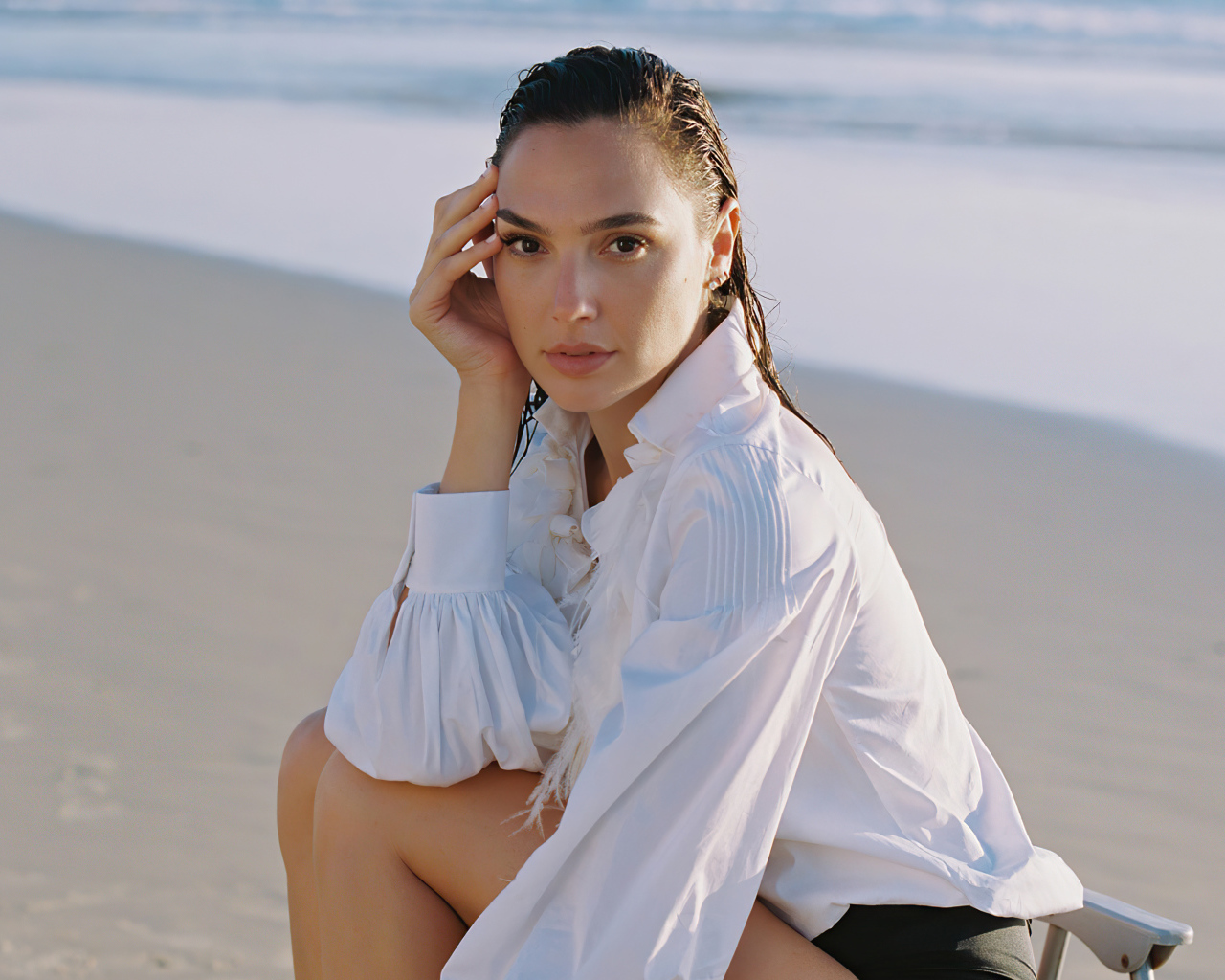 Актриса Галь Гадот в белой рубашке сидит на песке