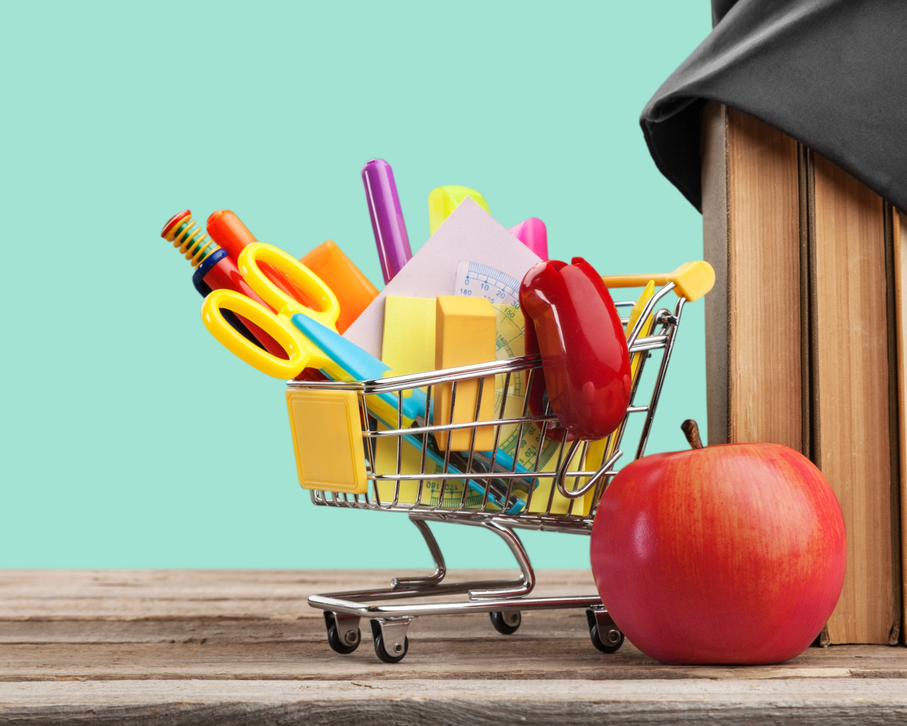 Канцелярские предметы в тележке на столе с яблоком и книгами 