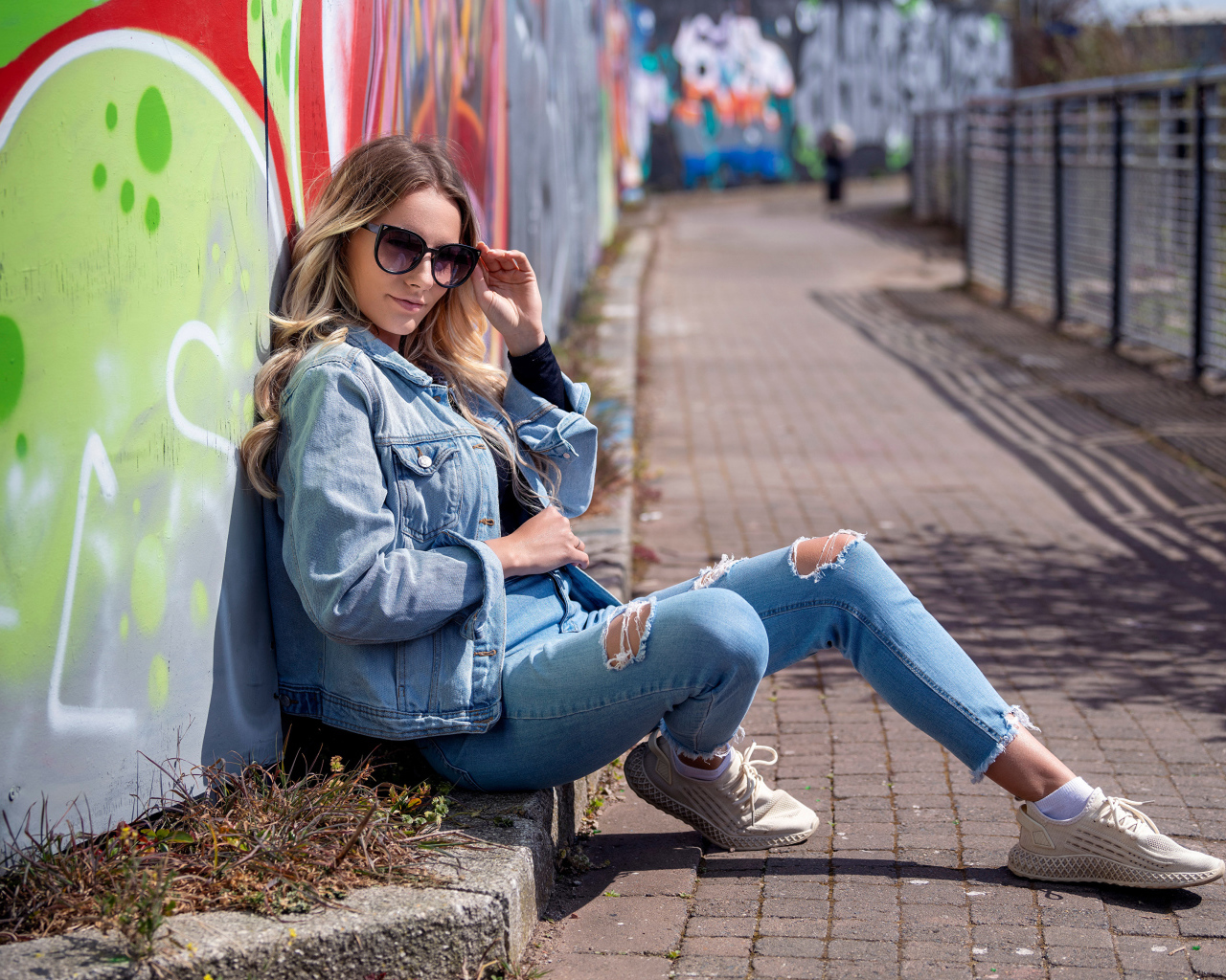 Красивая девушка в джинсовом костюме сидит у стены 