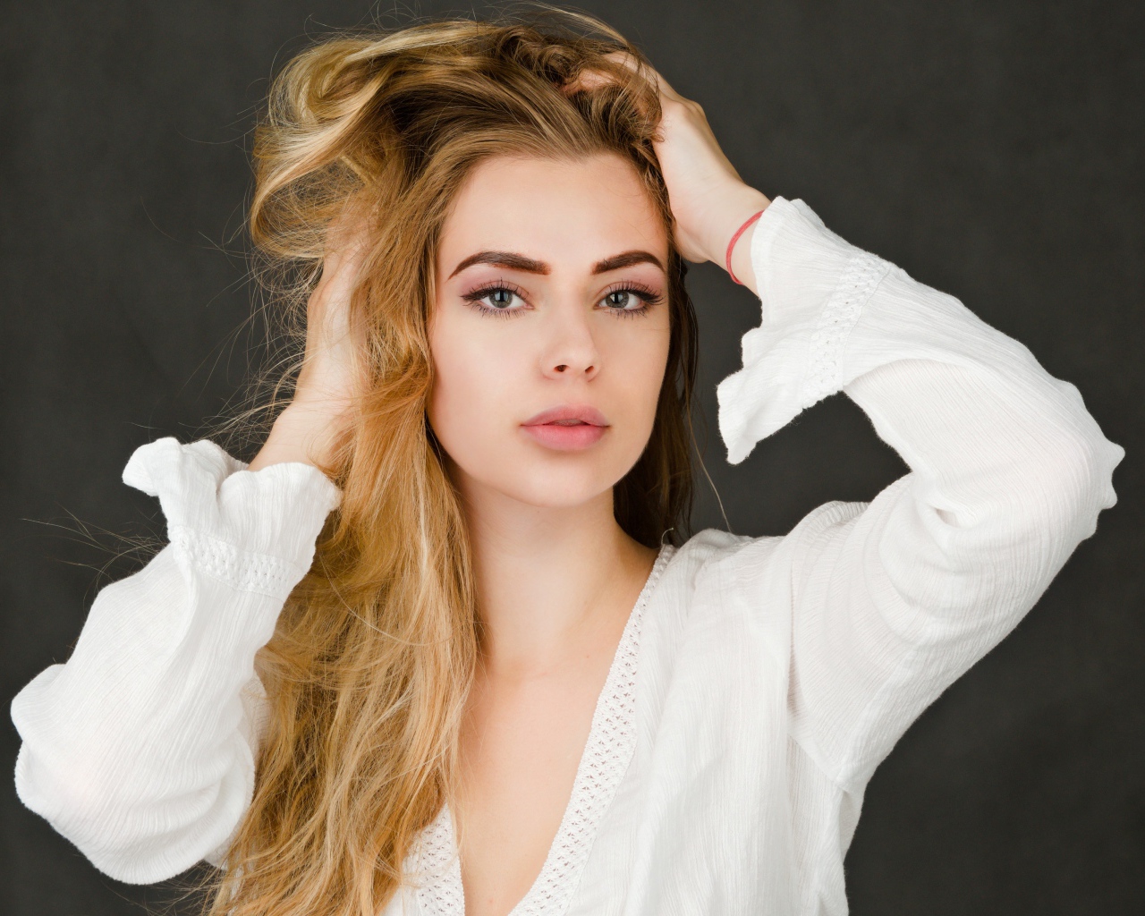 Блондинка в белой кофте с руками в голове на сером фоне 