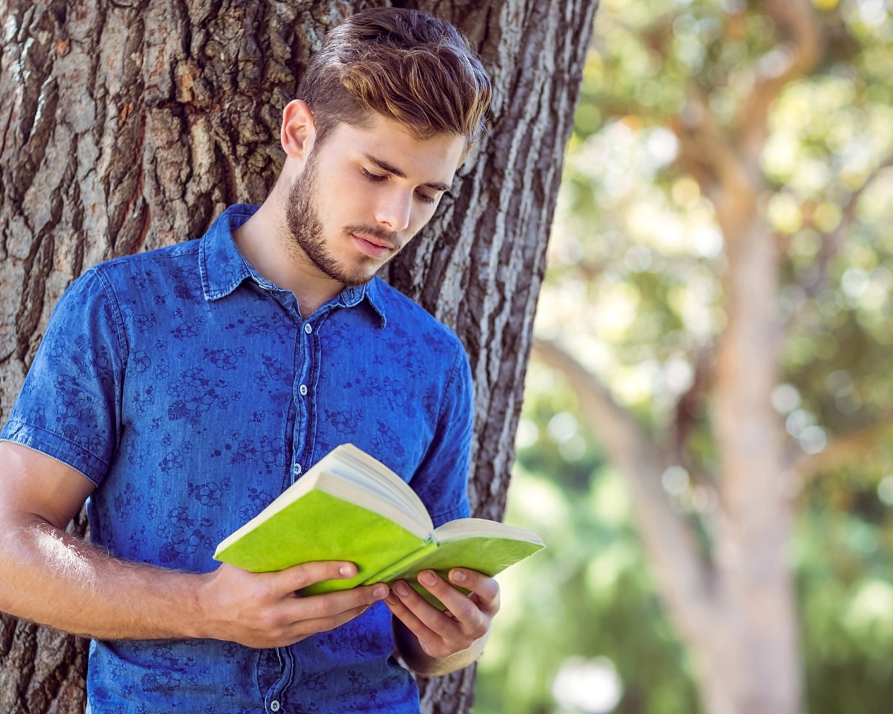 Красивый молодой мужчина стоит с книгой под деревом 