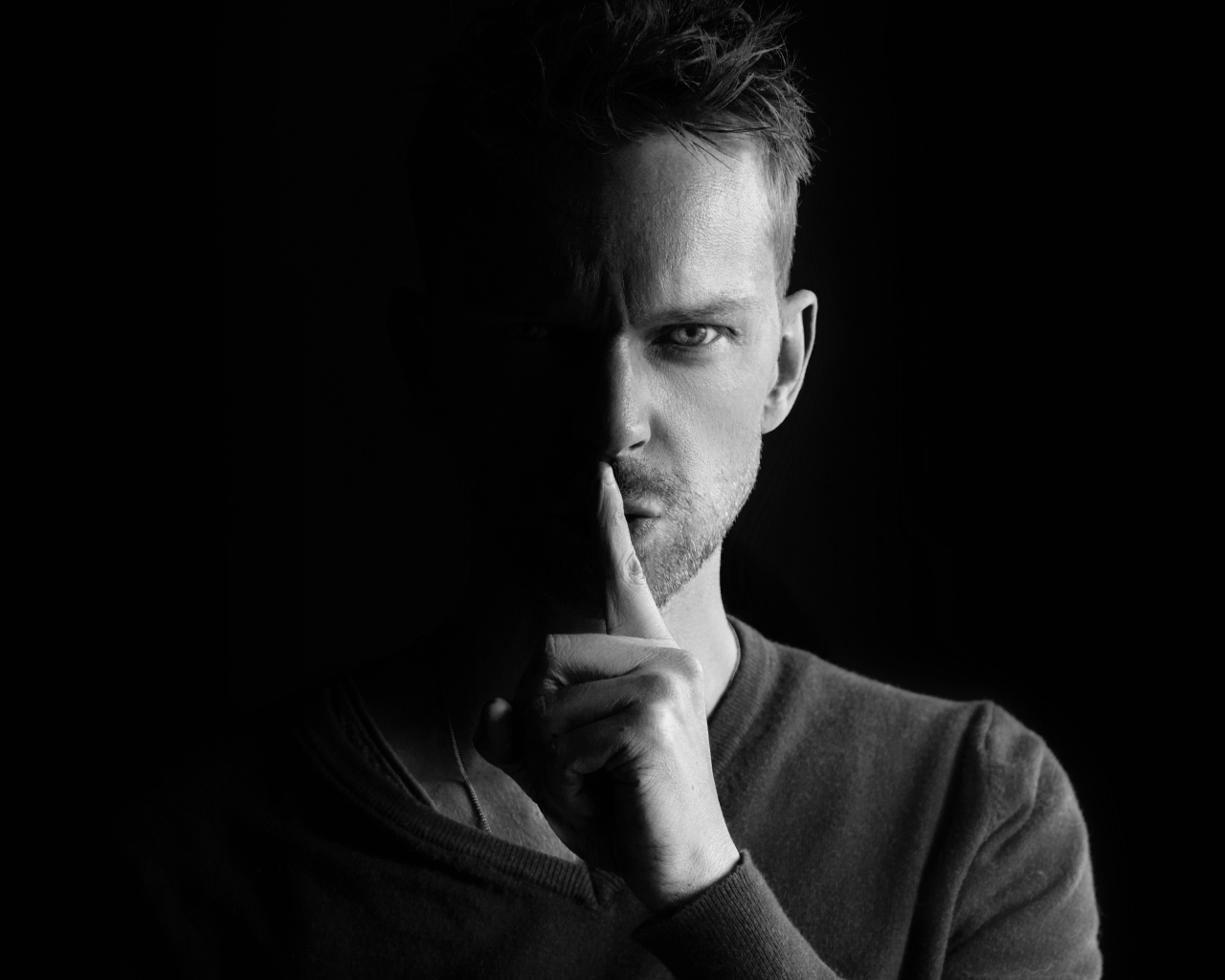 Молодой мужчина закрыл рот пальцем на черном фоне 