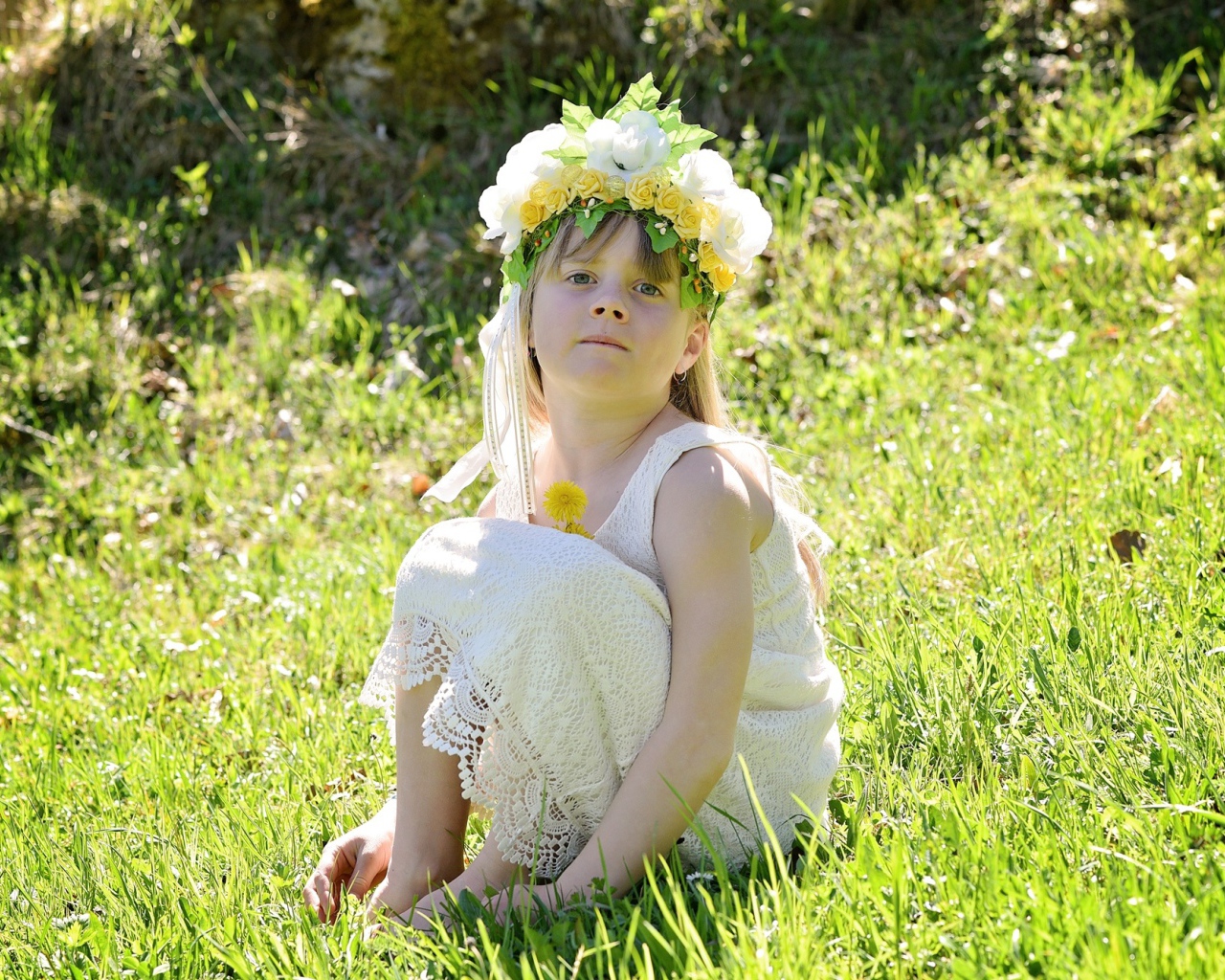 Маленькая девочка с венком на голове сидит на зеленой траве