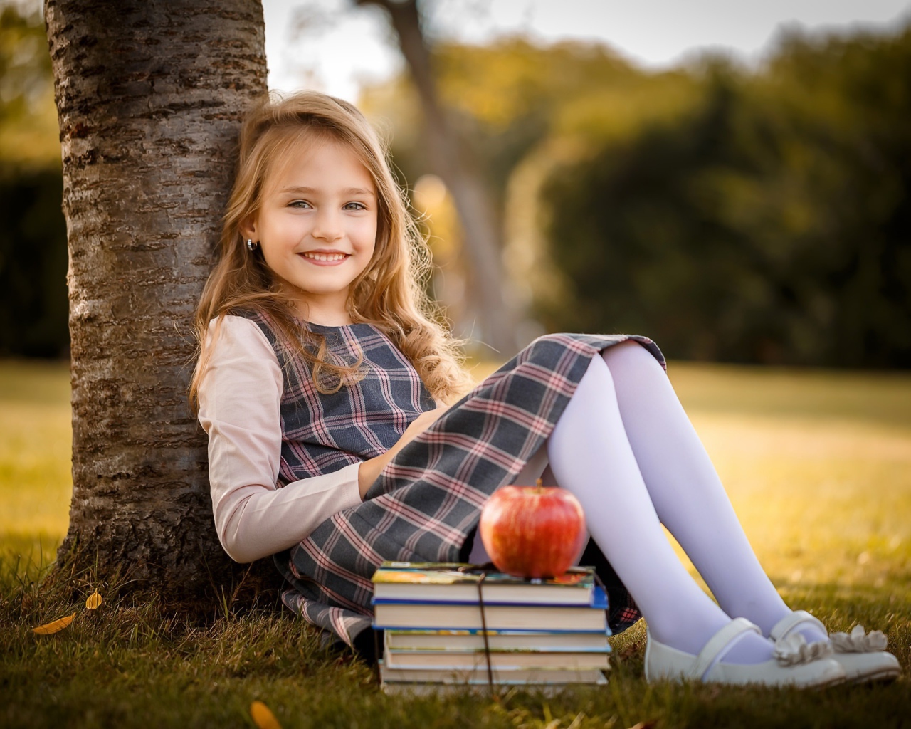 Маленькая девочка с книгами и яблоком сидит под деревом 