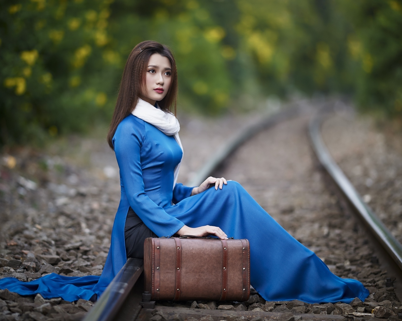 Красивая девушка азиатка сидит на рельсах с чемоданом