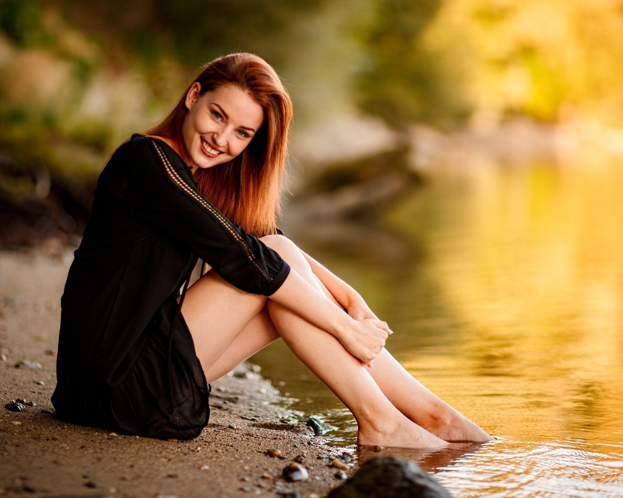 Улыбающаяся девушка в черном платье сидит у воды