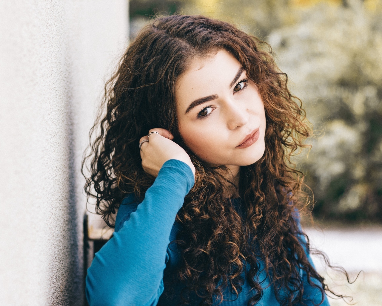 Кареглазая девушка с кудрявыми волосами стоит у стены