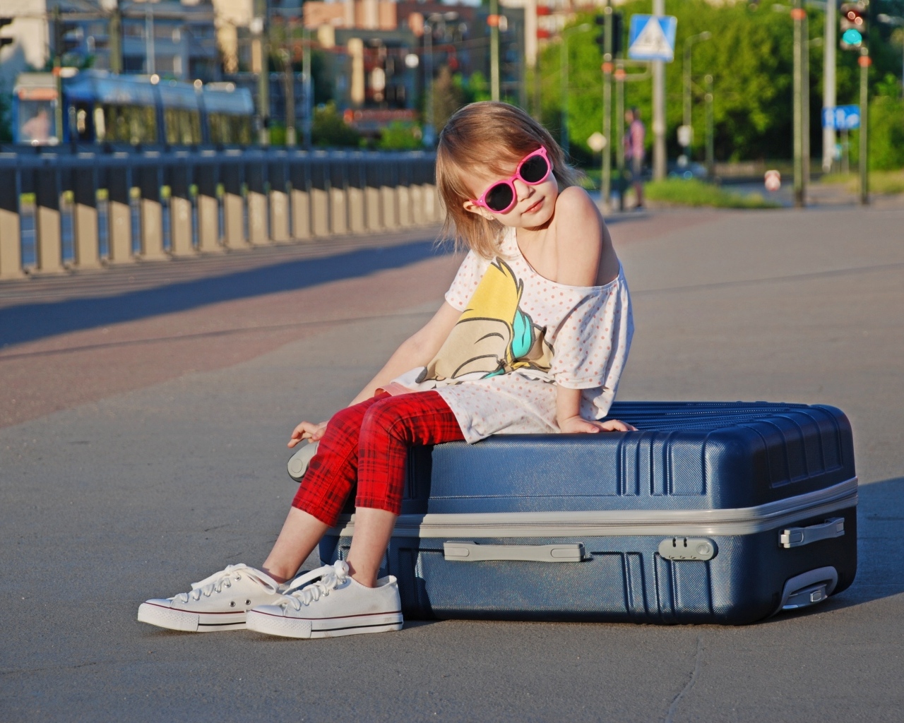 Девочка в солнечных очках сидит на чемодане на дороге