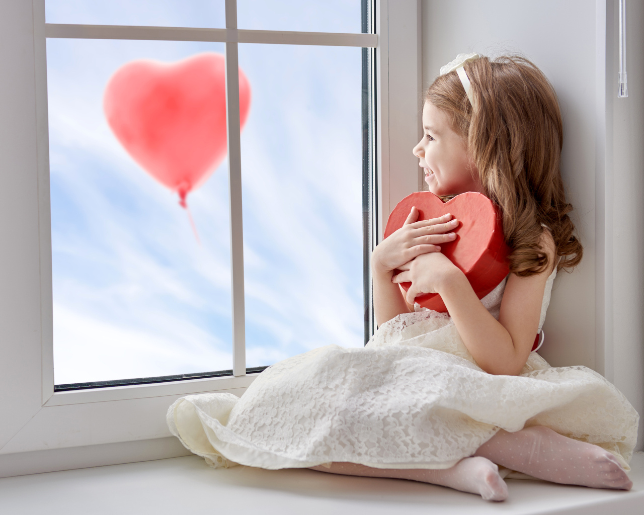 Маленькая девочка с коробкой в форме сердца сидит у окна