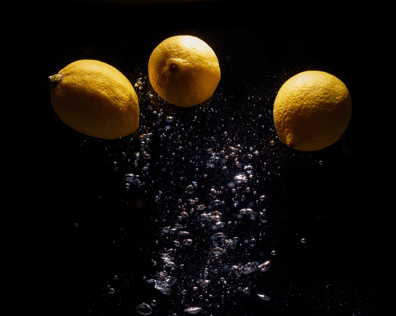 Три желтых лимона с пузырями в воде на черном фоне