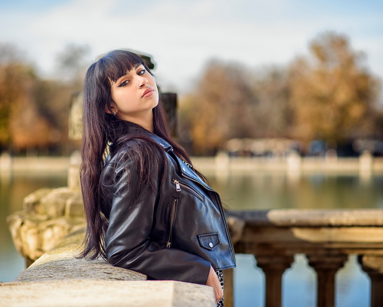 Девушка в черной куртке стоит на мосту у воды