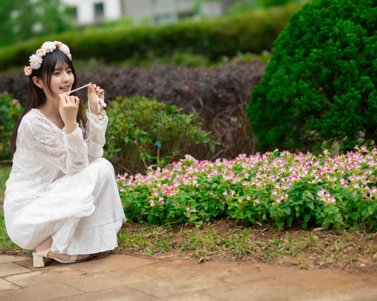 Милая азиатка в белом наряде сидит у клумбы с цветами