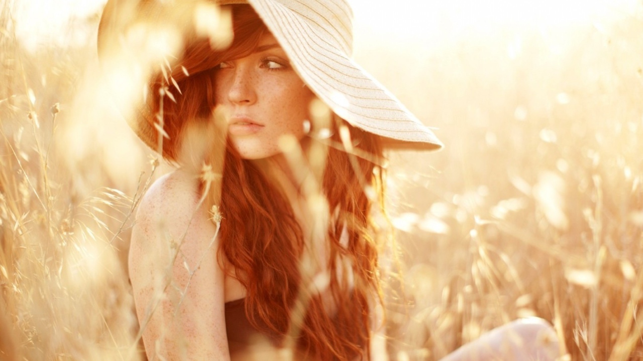 Рыжая девушка в шляпе сидит среди травы на поле