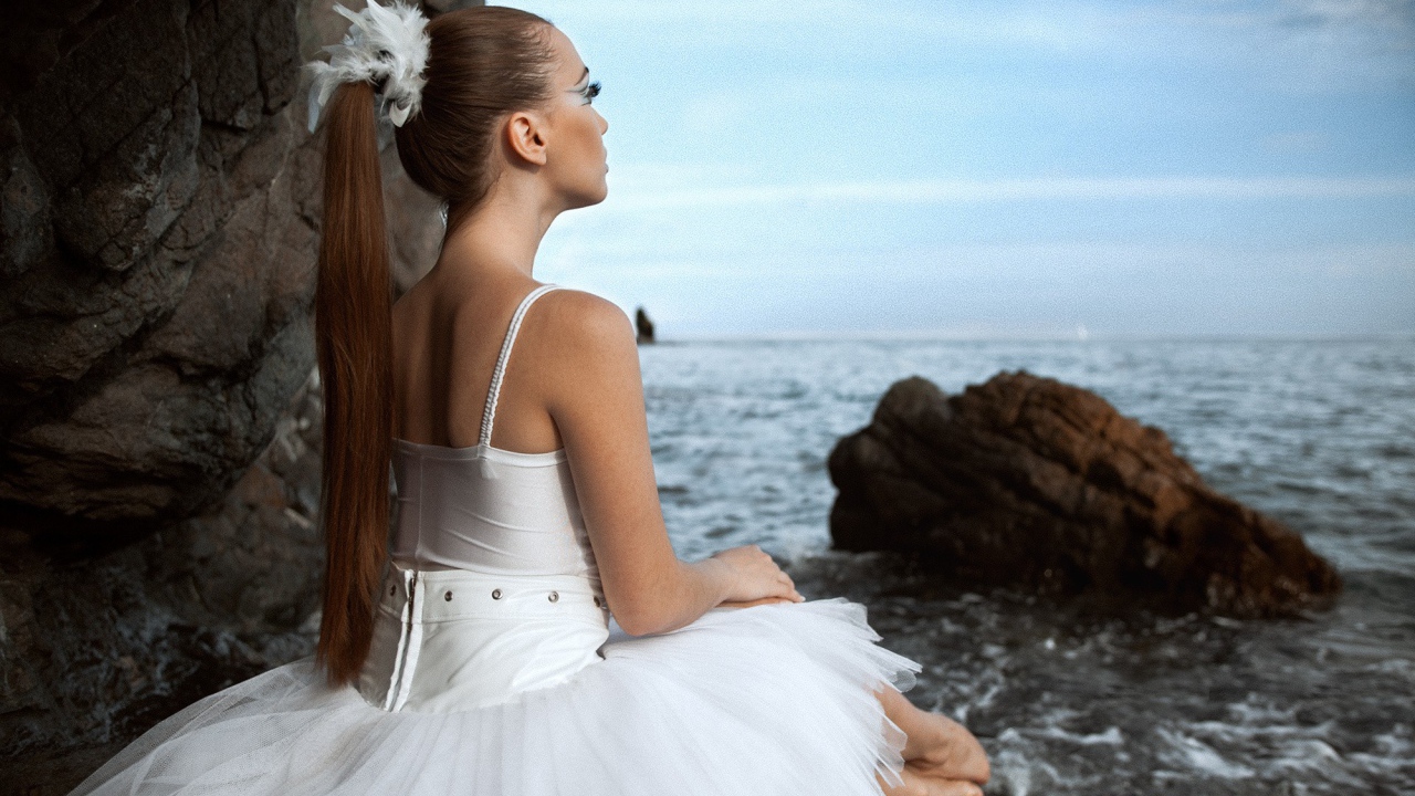Балерина в белом платье сидит на берегу моря