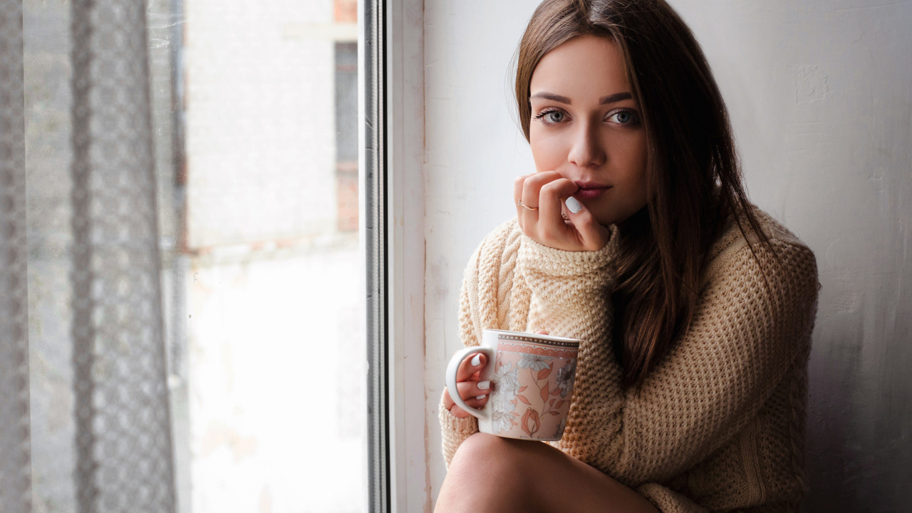 Молодая девушка в свитере сидит с чашкой на подоконнике