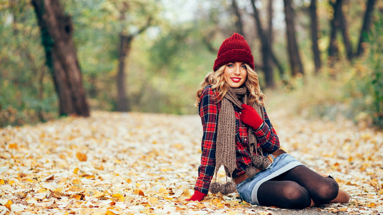 Молодая улыбающаяся девушка в красной шапке сидит на листве осенью