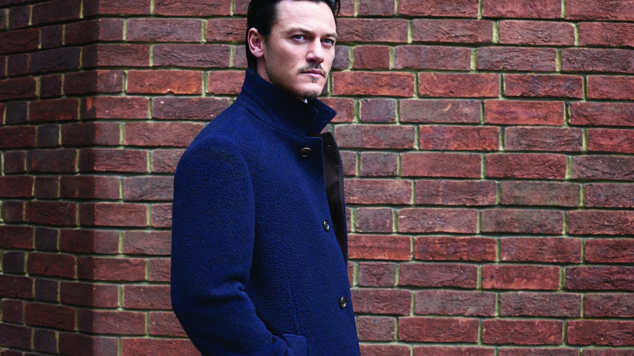 Актер Люк Эванс в синем плаще у кирпичной стены