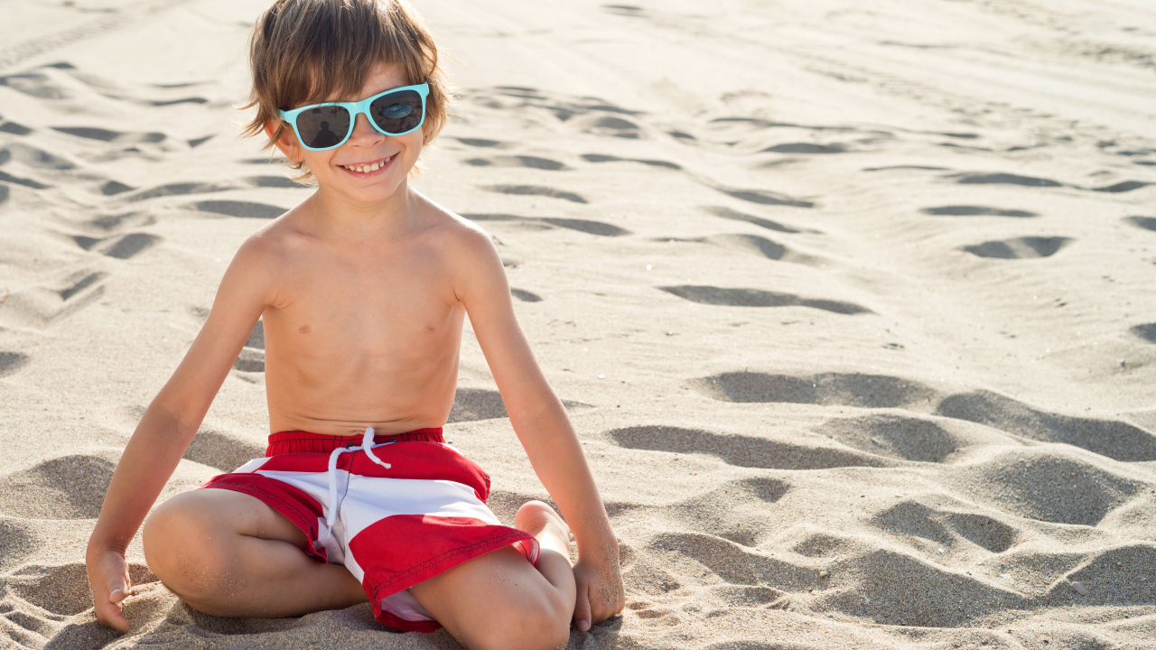 Маленький улыбающийся мальчик в очках сидит на песке