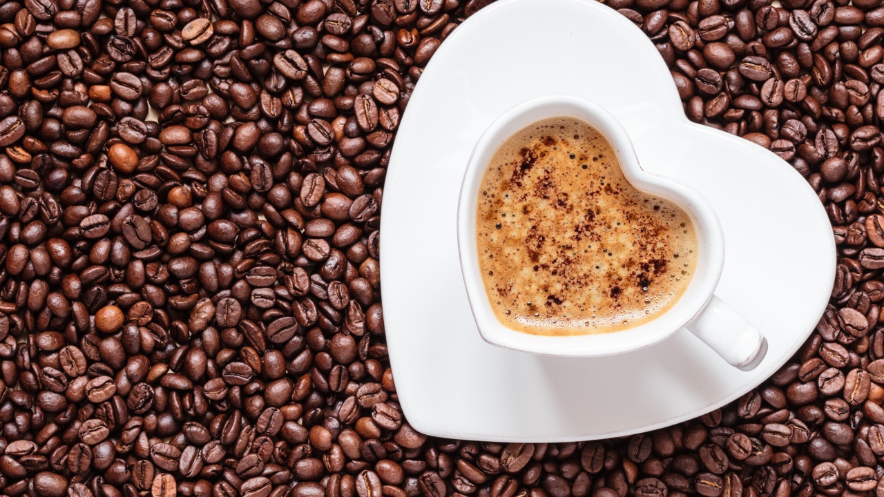 Белая чашка кофе в форме сердца стоит на кофейных зернах