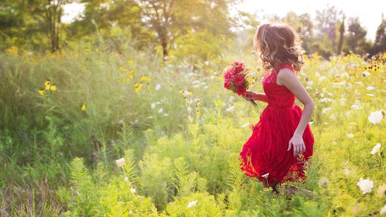 Красивая девушка в красном платье гуляет с букетом в руках по траве