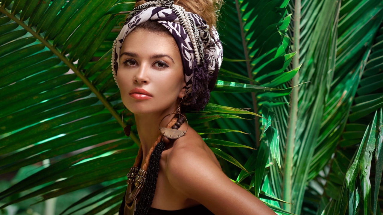 Красивая женщина в тюрбане с украшениями среди зеленых пальмовых листьев