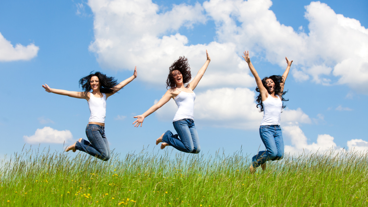 Три молодые девушки прыгают в зеленой траве на фоне неба 