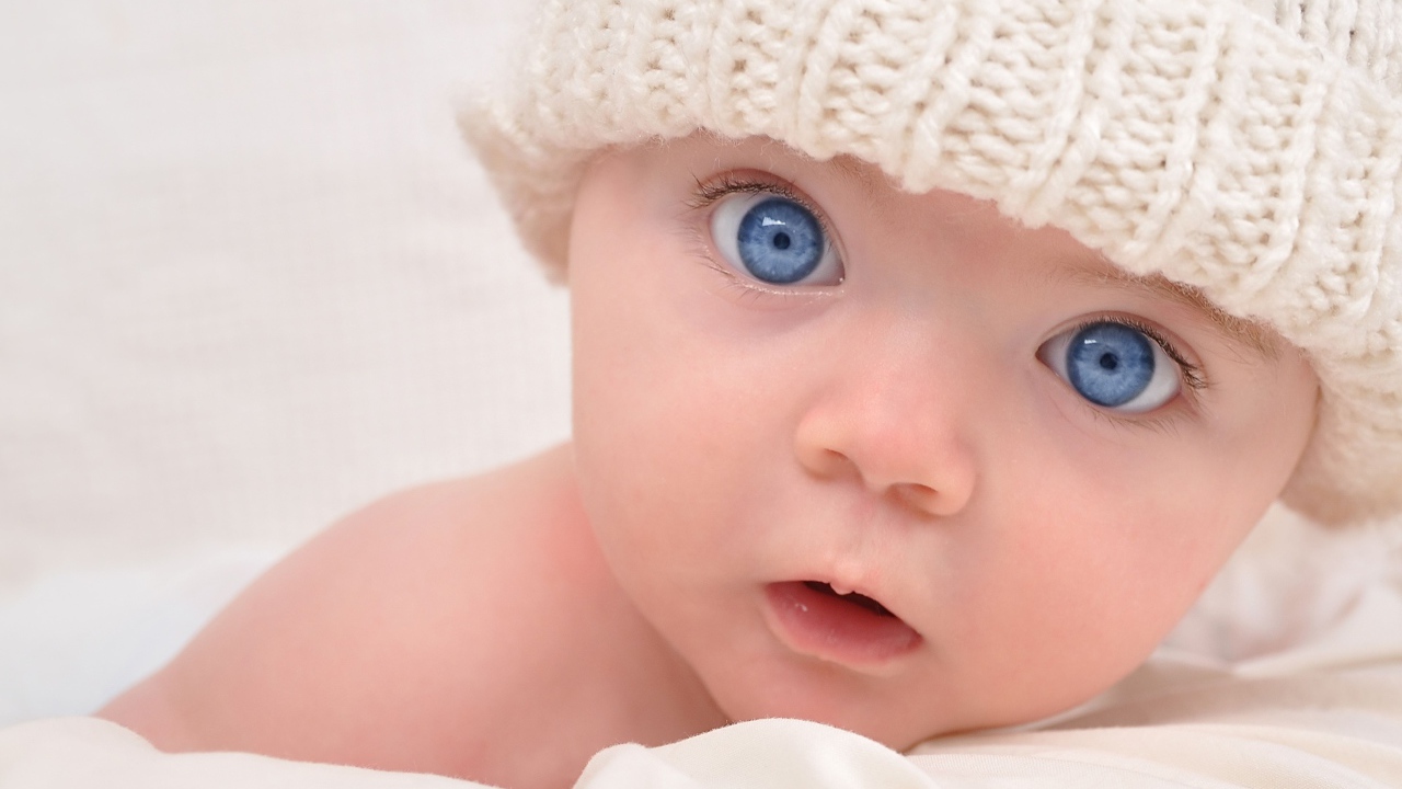 Голубоглазый малыш в белой вязаной шапке на сером фоне