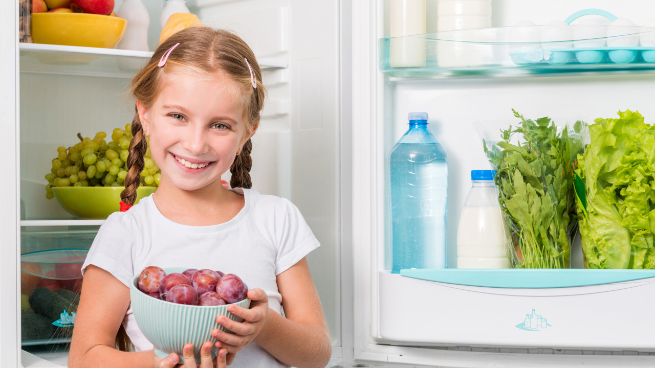 Маленькая девочка с миской со сливами в руках у холодильника 