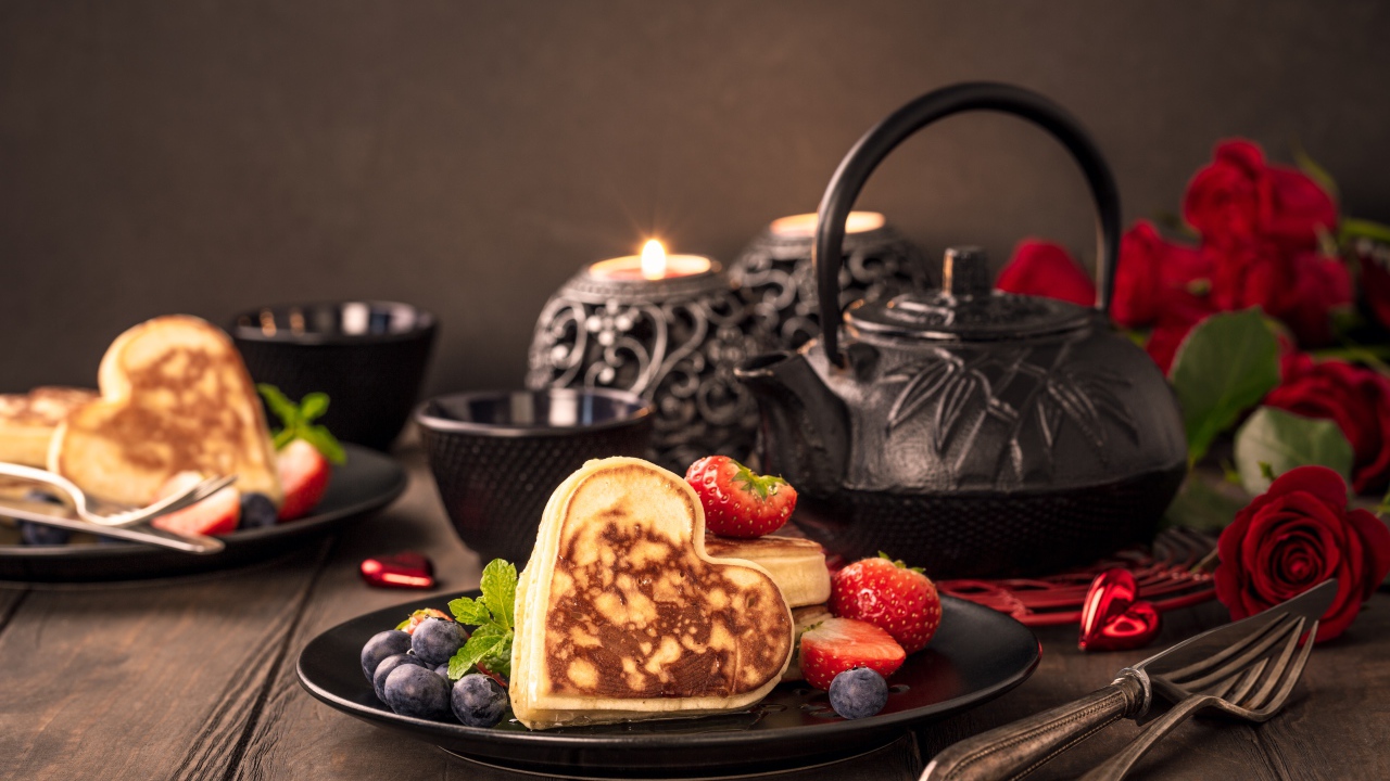 Оладьи в форме сердца с ягодами черники и клубники на столе с чаем и свечами