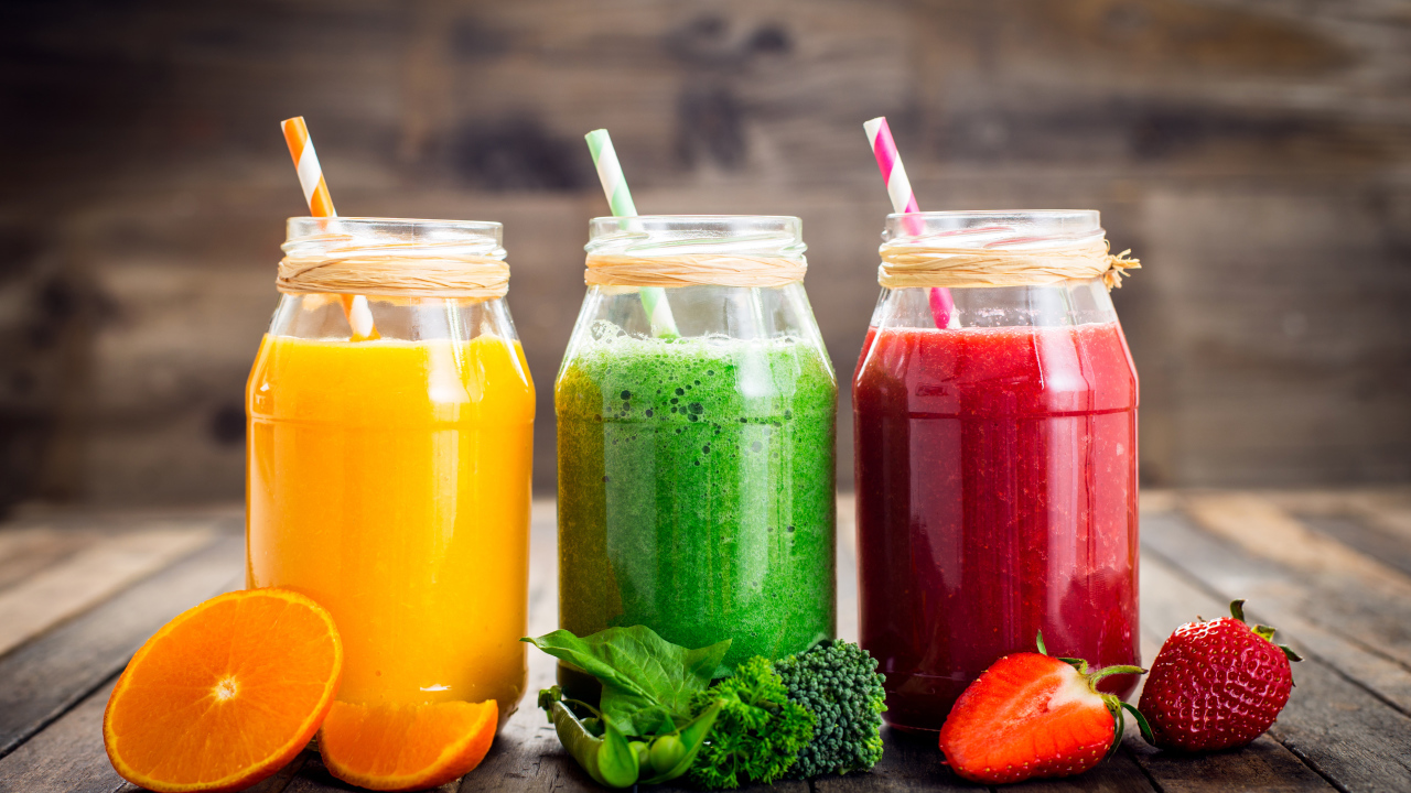 Три банки смузи с трубочкой на столе с апельсином, брокколи и клубникой
