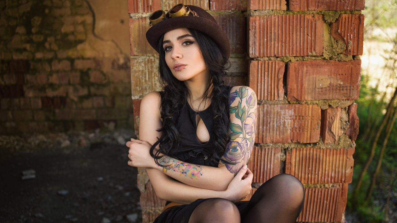 Девушка в шляпе с татуировками на руке сидит у стены