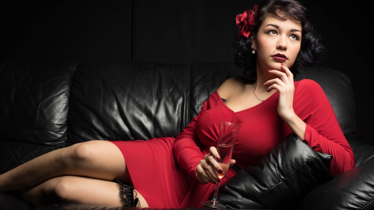 Девушка в красном платье с бокалом в руке лежит на кожаном диване