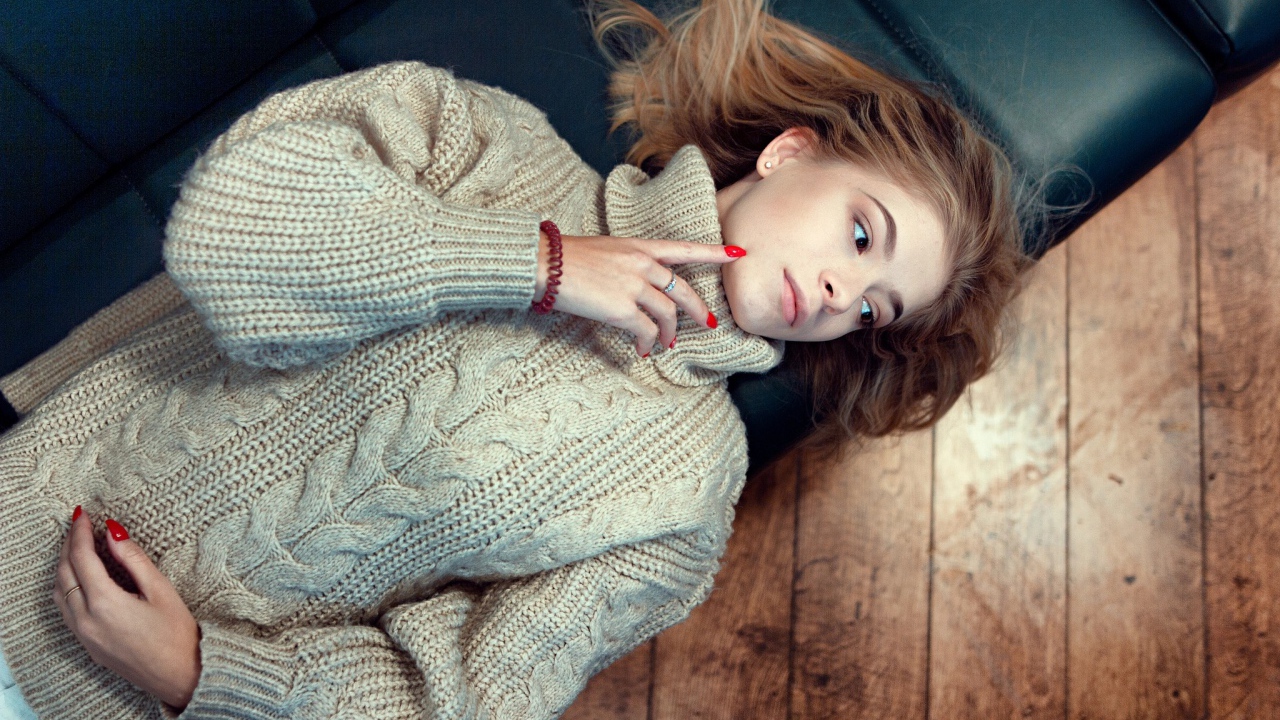 Молодая девушка в вязаном свитере лежит на черном кожаном диване