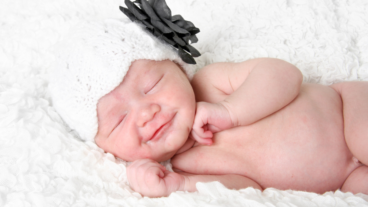 Улыбающийся спящий ребенок в белой вязаной шапке с цветком