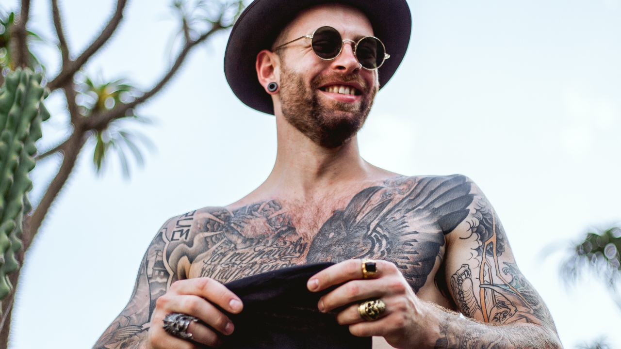 Мужчина с татуировками на теле в черной шляпе и очках
