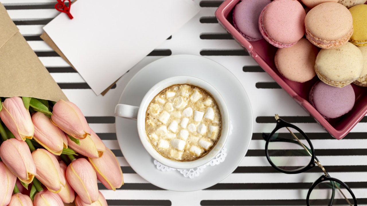 Чашка какао с маршмеллоу на столе с букетом тюльпанов, десертом макарун и очками