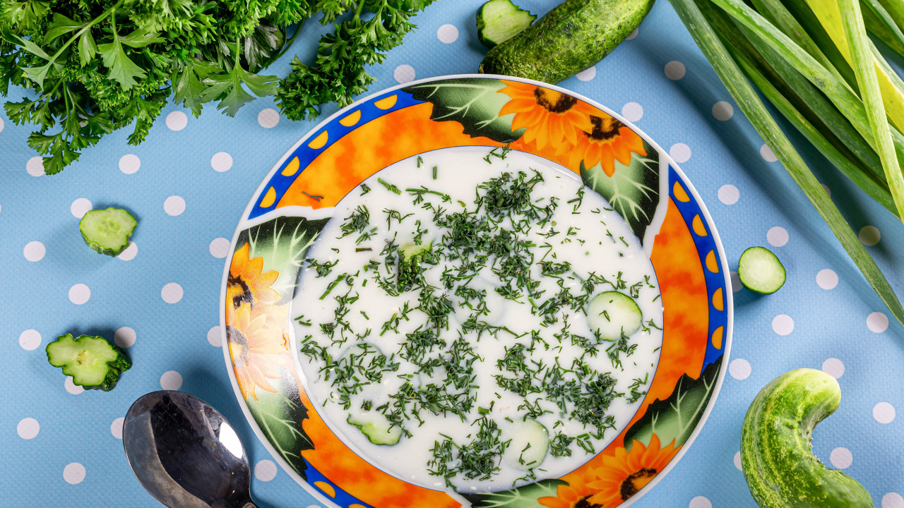 Окрошка в тарелке на столе с зеленью и огурцами