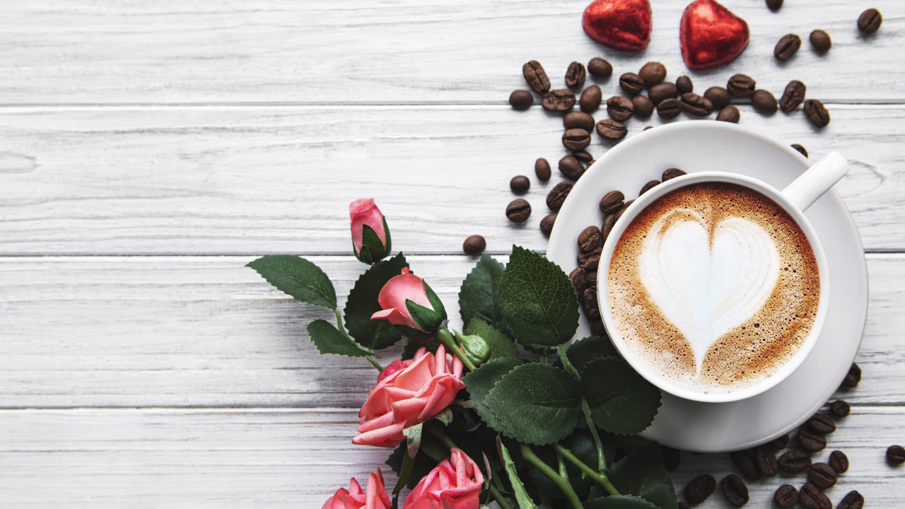 Чашка кофе с рисунком на пене на столе с букетом роз, зернами и конфетами