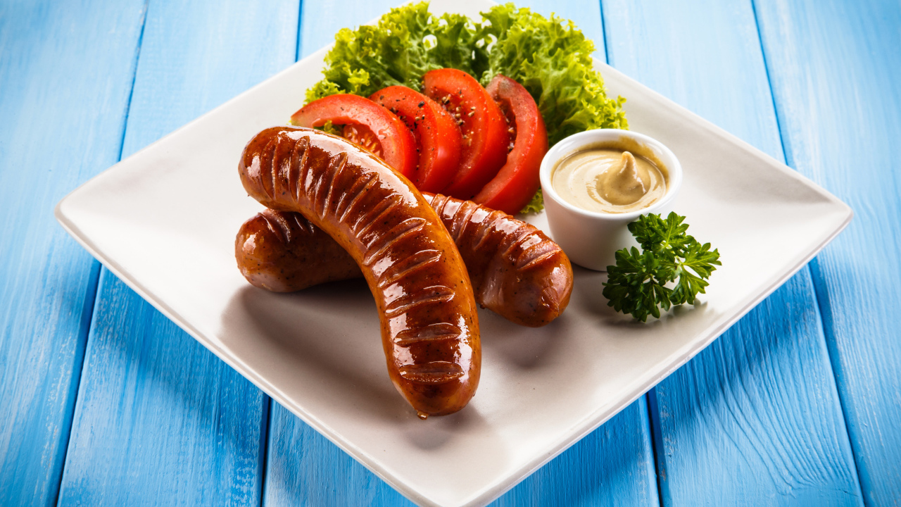 Две жареные колбаски на тарелке с помидорами, горчицей и листьями салата 