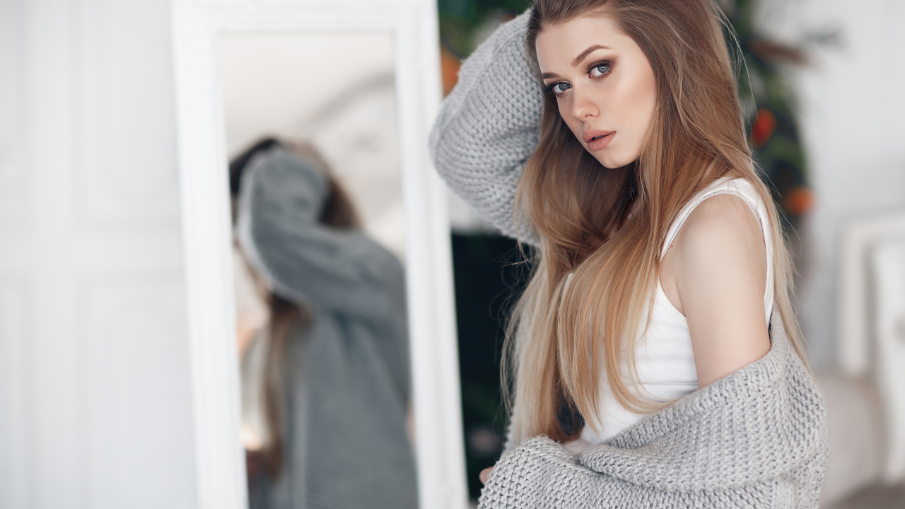 Молодая девушка в серой кофте стоит у зеркала 