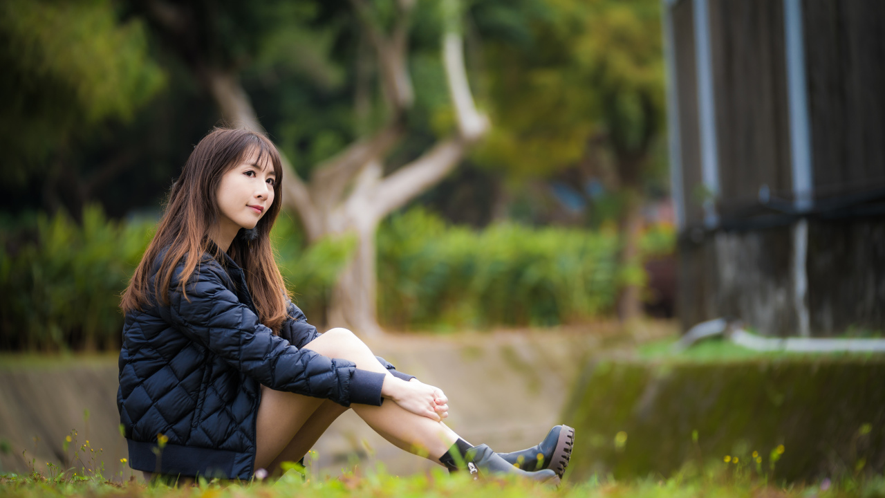 Девушка азиатка в черной куртке сидит на траве