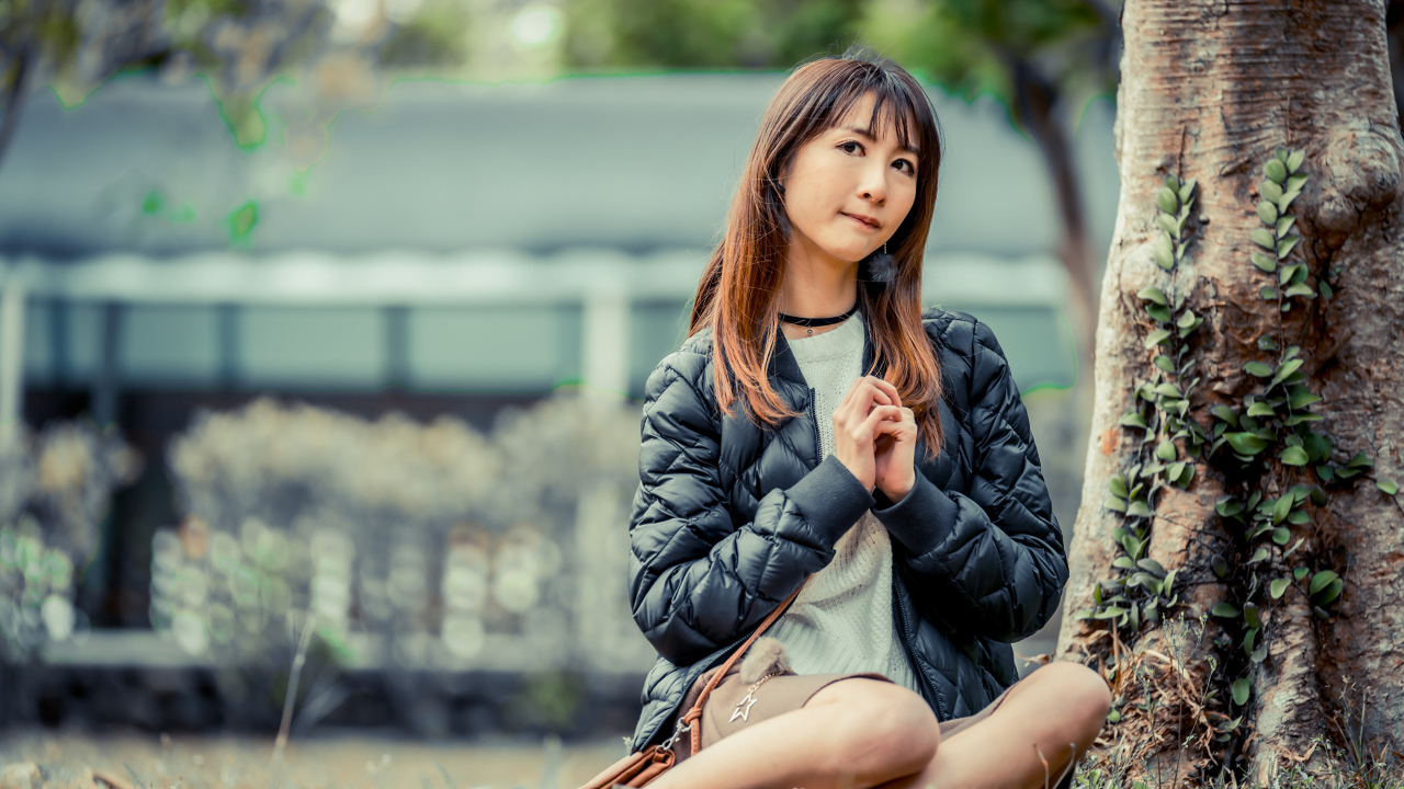 Девушка азиатка в черной куртке сидит под деревом