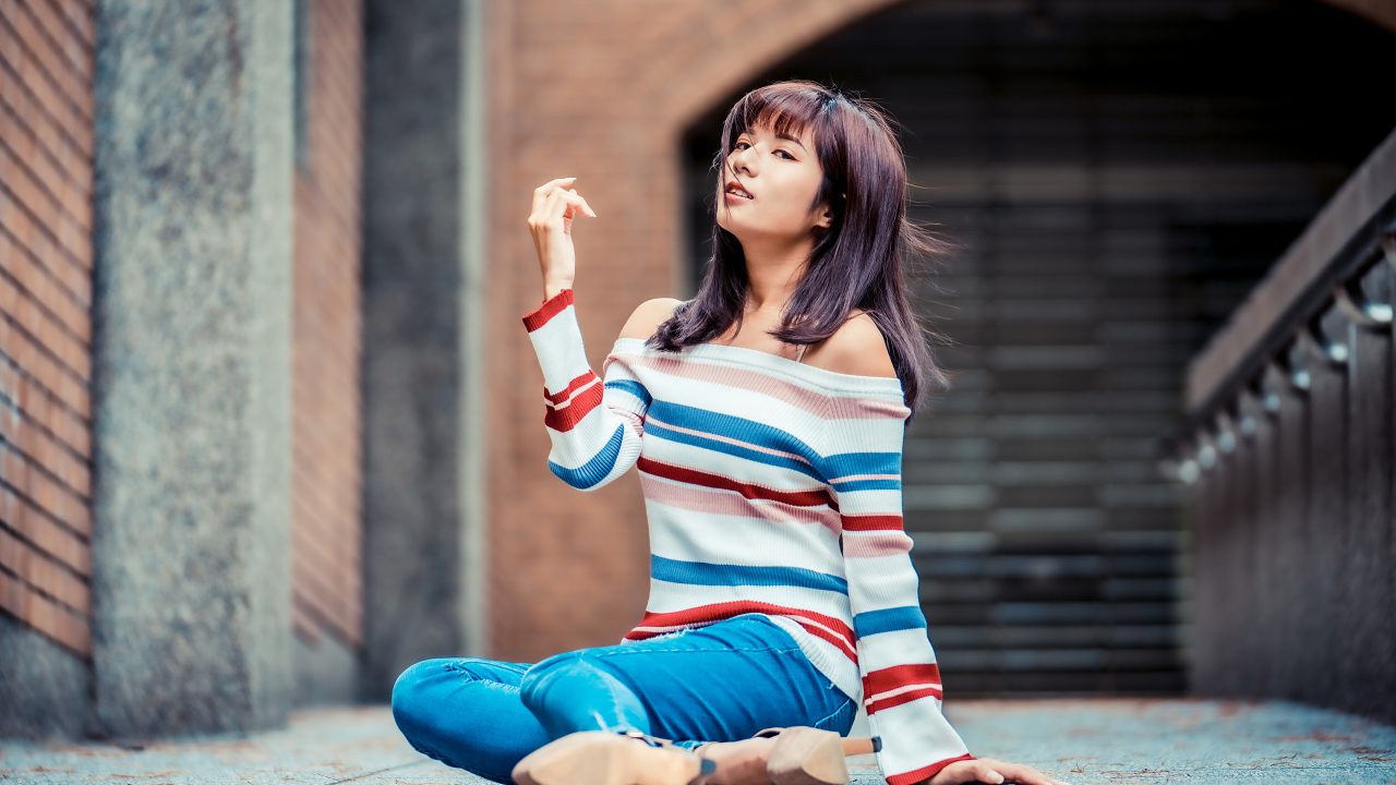 Девушка азиатка в джинсах и полосатом свитере сидит на асфальте