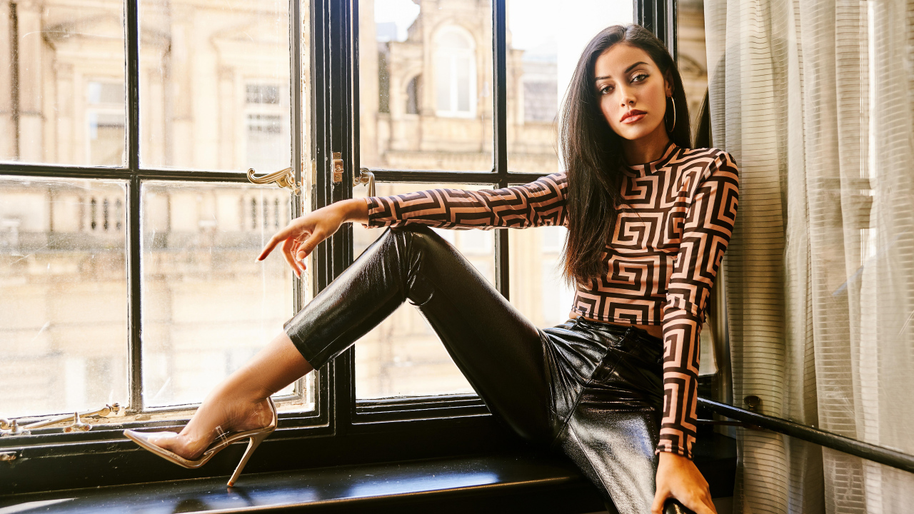 Красивая брюнетка в черных брюках сидит на окне 