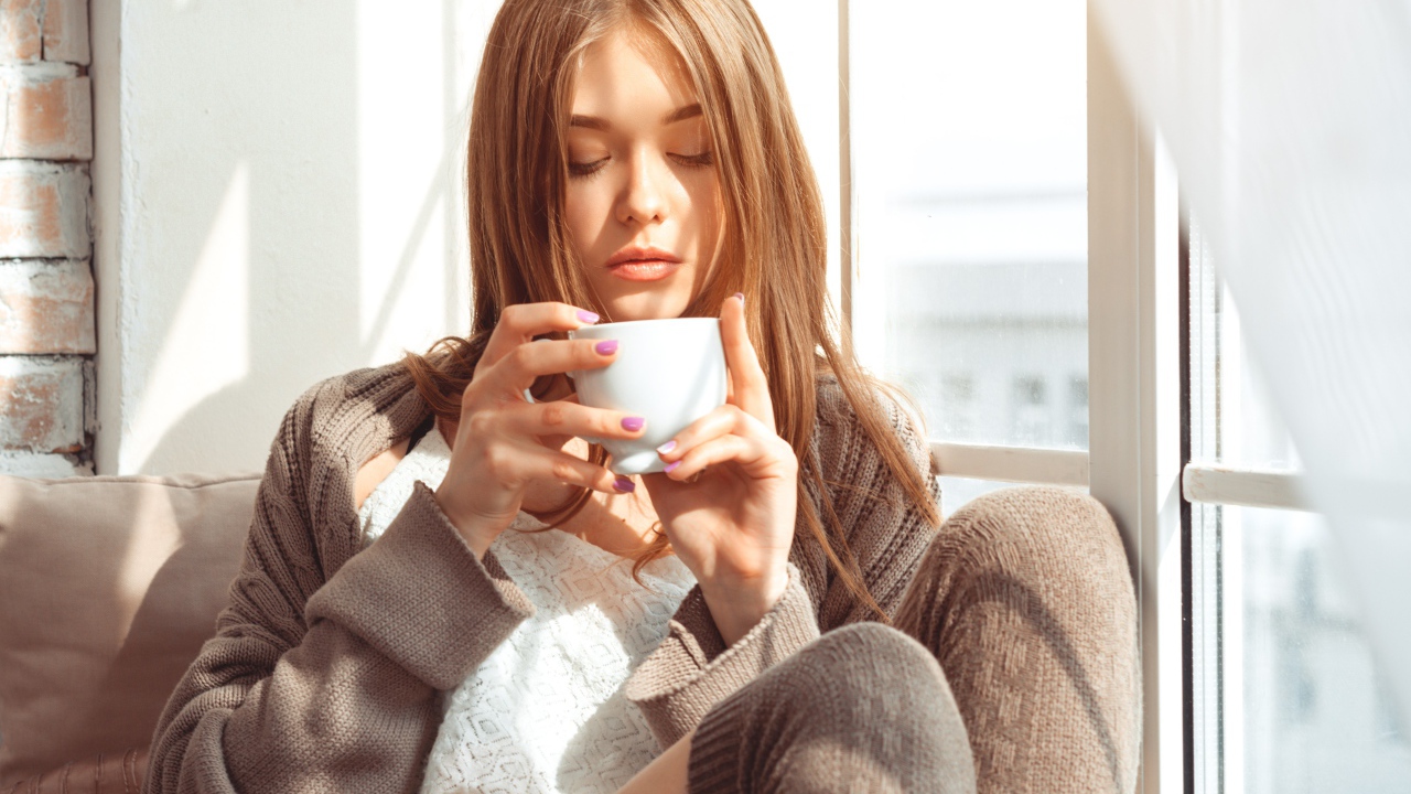 Красивая девушка в свитере пьет кофе у окна