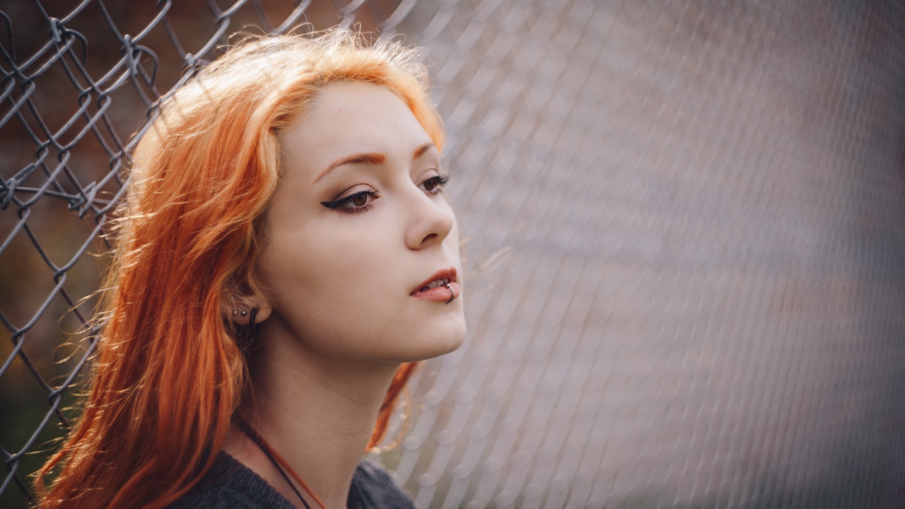 Красивая рыжеволосая девушка с пирсингом у железной сетки 