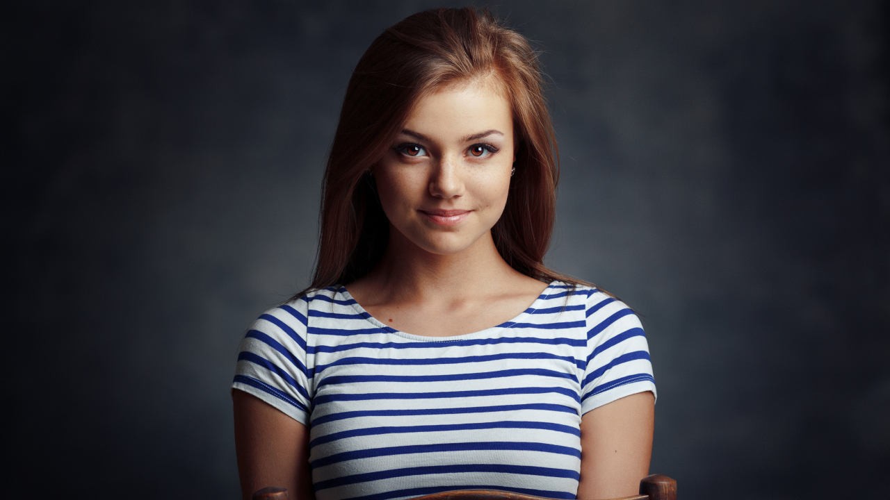 Милая кареглазая девушка в полосатой майке сидит на стуле 